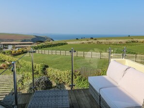 A deck with a white sofa and a view of a grassy field with cows, a wooden fence, and a building in the background. - Coastman's Nest (Newquay)