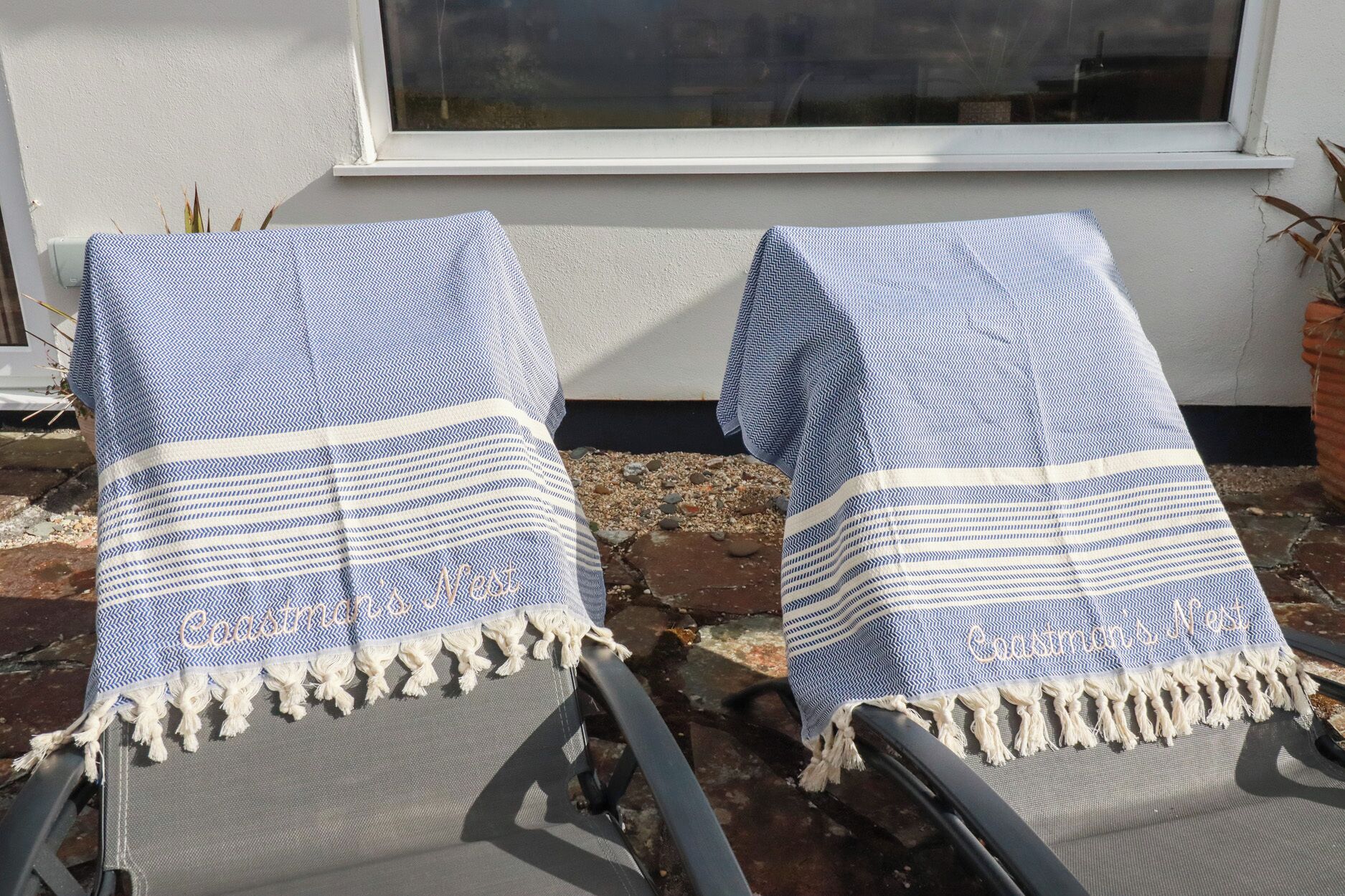 Two outdoor lounge chairs covered with patterned blankets, positioned in front of a white building with a window.