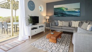 A modern living room with a grey sofa, a coffee table, a TV on a white stand, a wall clock, and a large ocean wave painting. - Coastman's Nest (Newquay)
