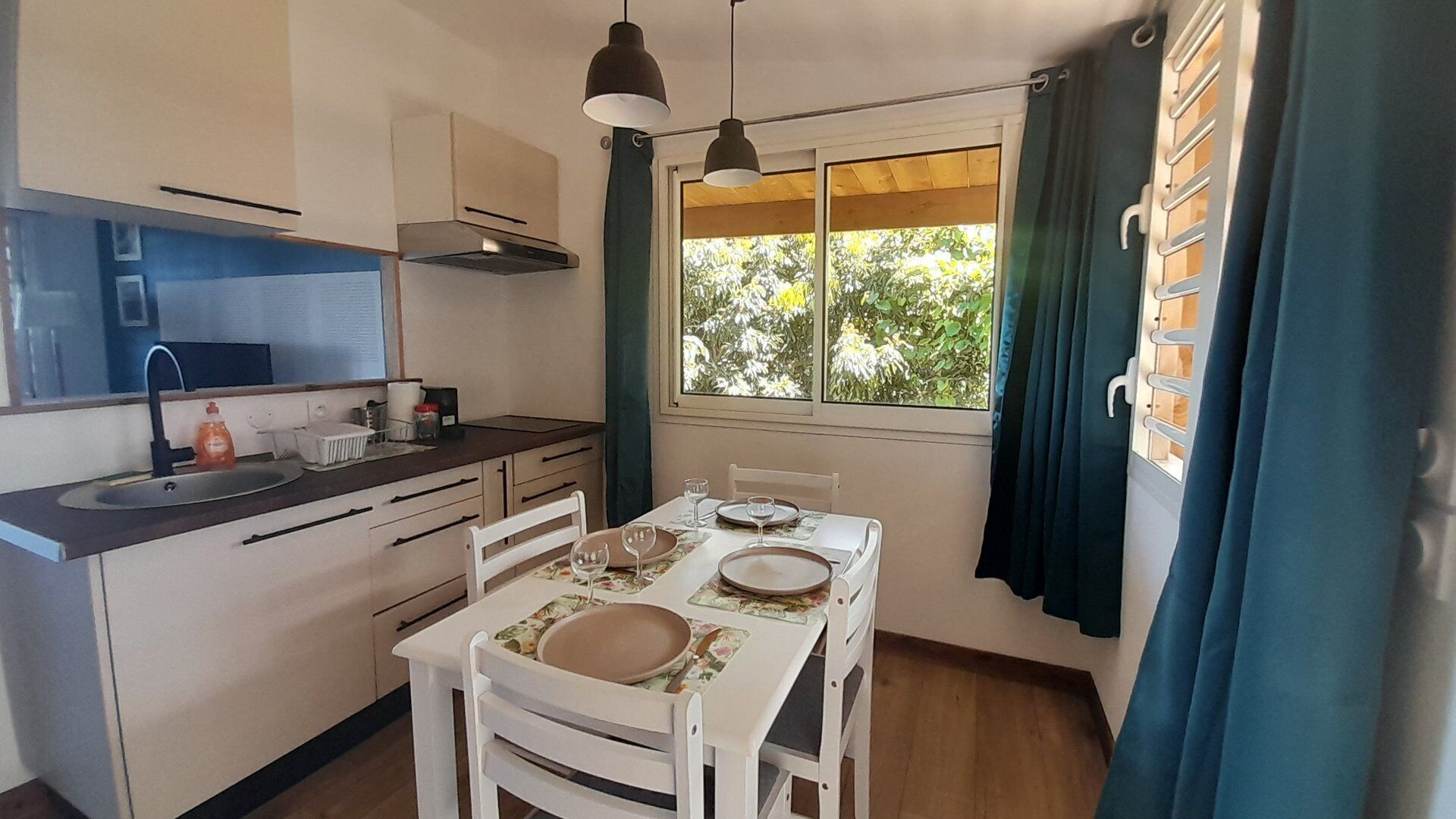 A compact kitchen with a dining area, a window with curtains, and a view of greenery outside.