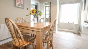 Un coin repas avec une table et des chaises en bois, un vase rempli de tournesols, un tableau encadré accroché au mur et une fenêtre avec des rideaux.