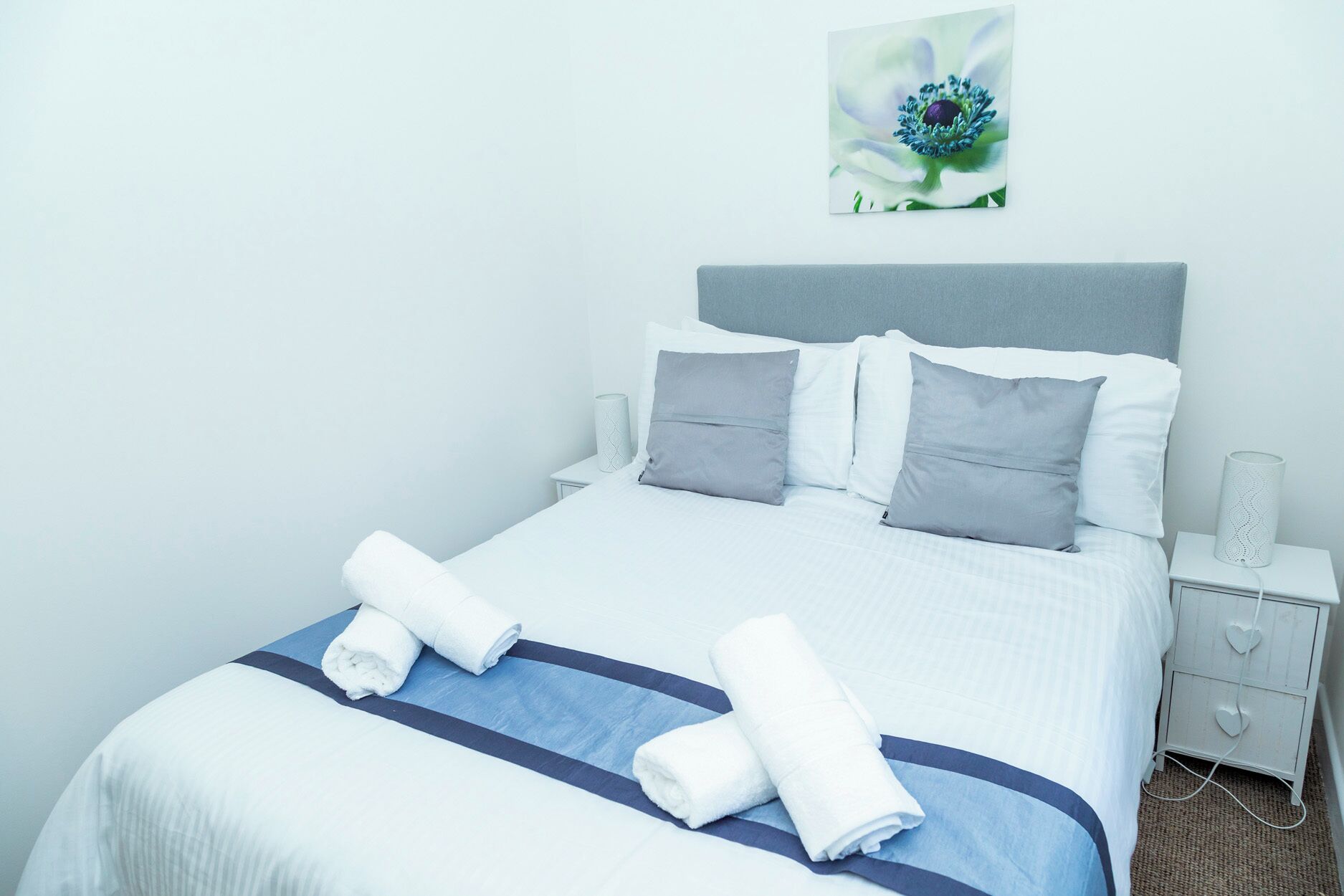 A neatly made bed with white bedding and a blue accent, two pillows, and two rolled towels on a blue accent strip.
