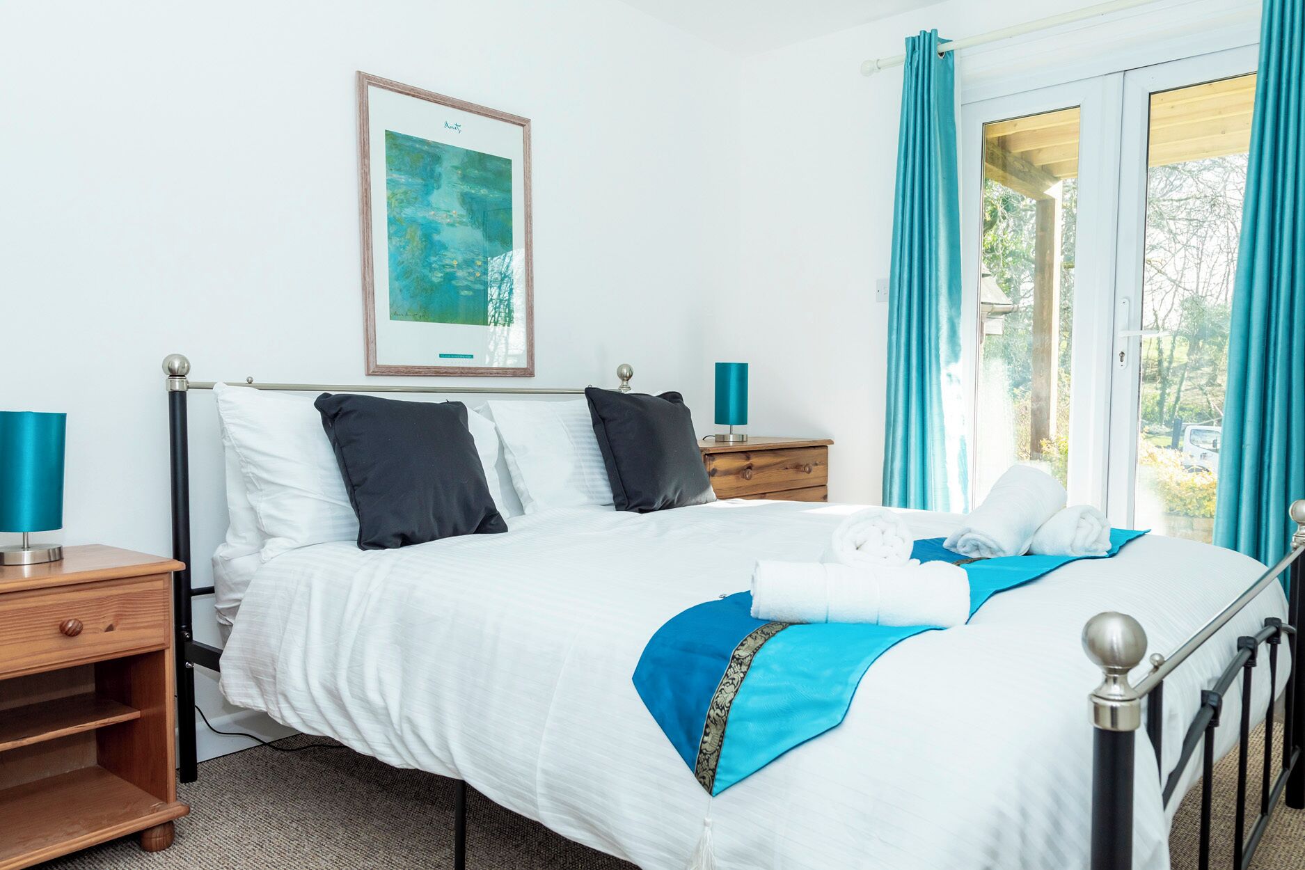 A neatly made bed with white and blue bedding, a framed artwork above it, and a view of greenery through the window.