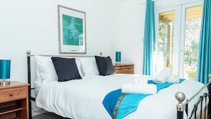 A neatly made bed with white and blue bedding, a framed artwork above it, and a view of greenery through the window. - Valley Lodge 40 (Gunnislake)