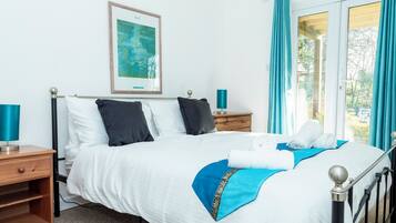 A neatly made bed with white and blue bedding, a framed artwork above it, and a view of greenery through the window.