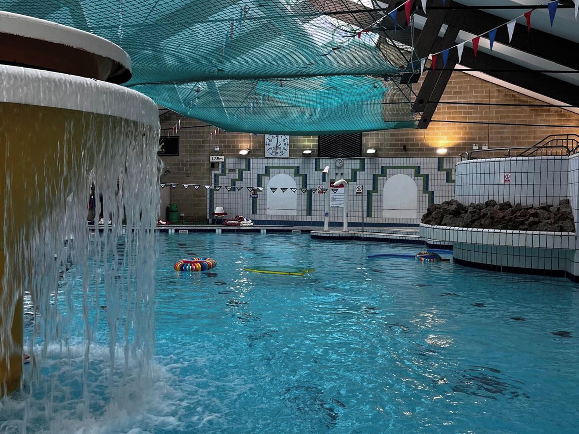 Ein Hallenbad mit Wasserfall, einer Uhr und einer Fliesenwand mit geometrischem Muster.