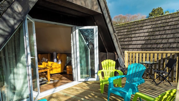 An outdoor deck with colorful Adirondack chairs, a wooden table, and a glass door leading to an interior space with a yellow sofa and a patterned rug. - Valley Lodge 40 (Gunnislake)