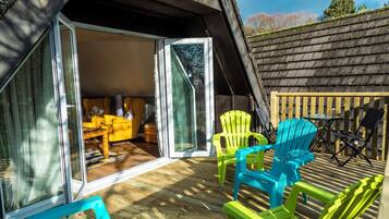 An outdoor deck with colorful Adirondack chairs, a wooden table, and a glass door leading to an interior space with a yellow sofa and a patterned rug.