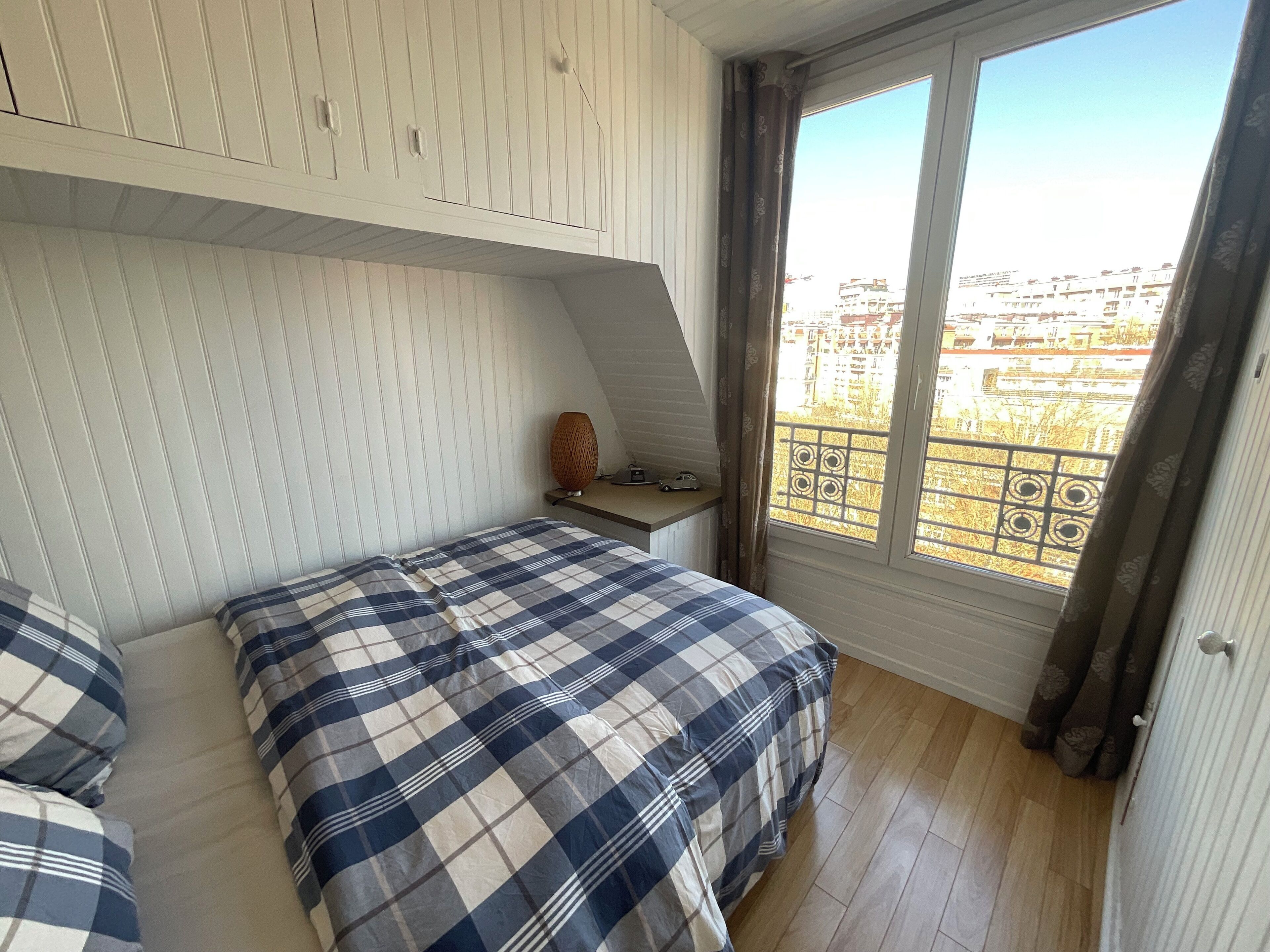 Un dormitorio con cama, mesita de noche, lámpara y ventana con vistas a edificios.