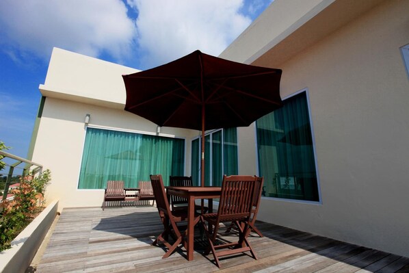 A modern outdoor patio with a wooden table and chairs, a large umbrella, and a glass door leading to a room with green curtains.