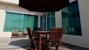 Een houten terras met een tafel en stoelen, een grote parasol, en een gebouw met grote ramen en gordijnen.