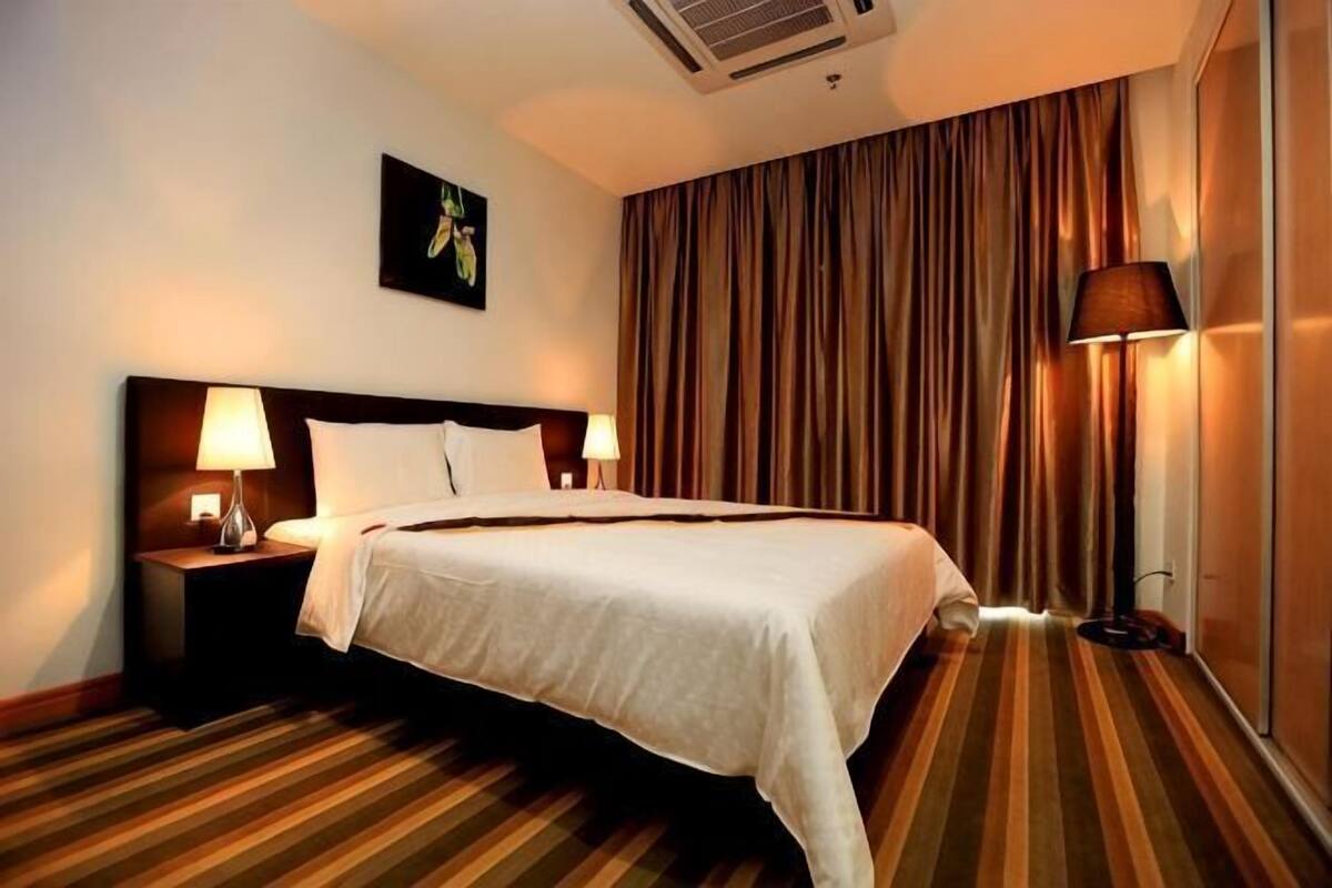Habitación de hotel con una cama grande, dos mesitas de noche con lámparas, un cabecero, una ventana con cortinas y un aire acondicionado instalado en la pared.