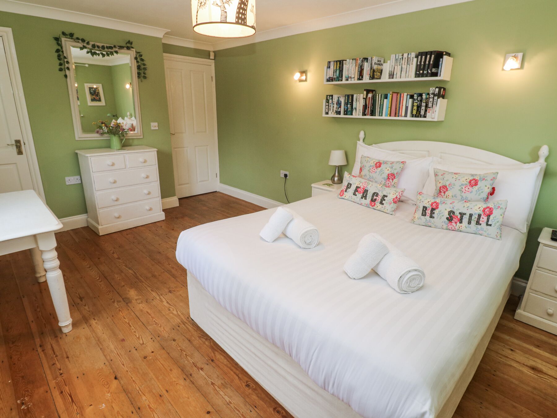 A bedroom with a large bed, a bedside table, a nightstand, a dresser, a mirror, and a bookshelf.