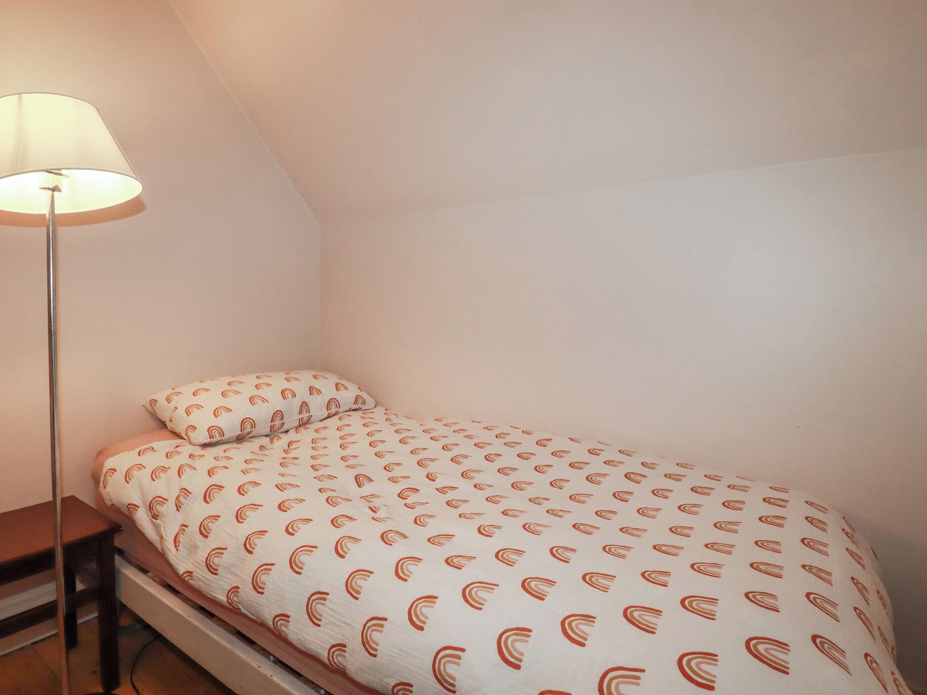 Une petite chambre avec un lit simple, un plafond en pente, une table de chevet et une lampe sur pied.