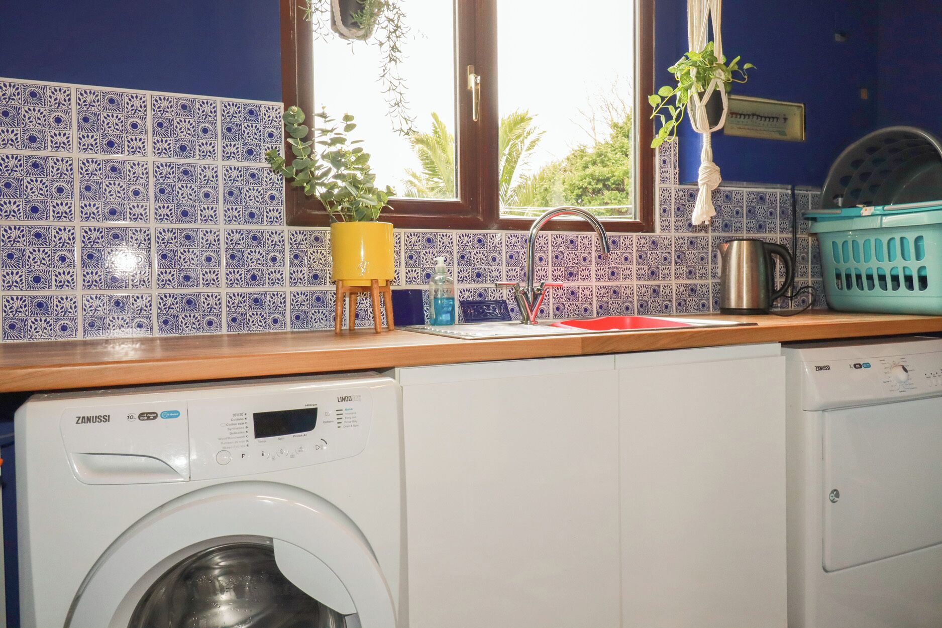 Une cuisine équipée d’un lave-linge, d’un évier et d’une fenêtre agrémentée de plantes.