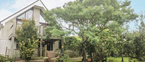 A house with a large tree in the front yard, a wooden deck, and a small porch with steps leading up to the entrance.