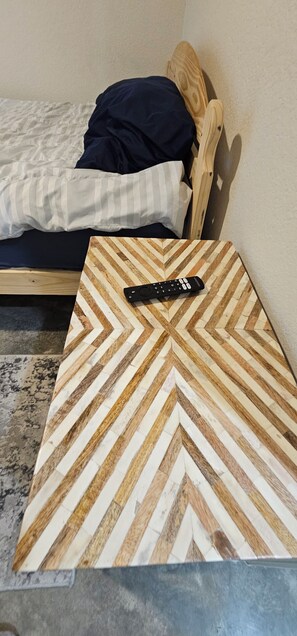 Una cama con cabecera de madera, una mesita de noche con un control remoto y una alfombra gris.