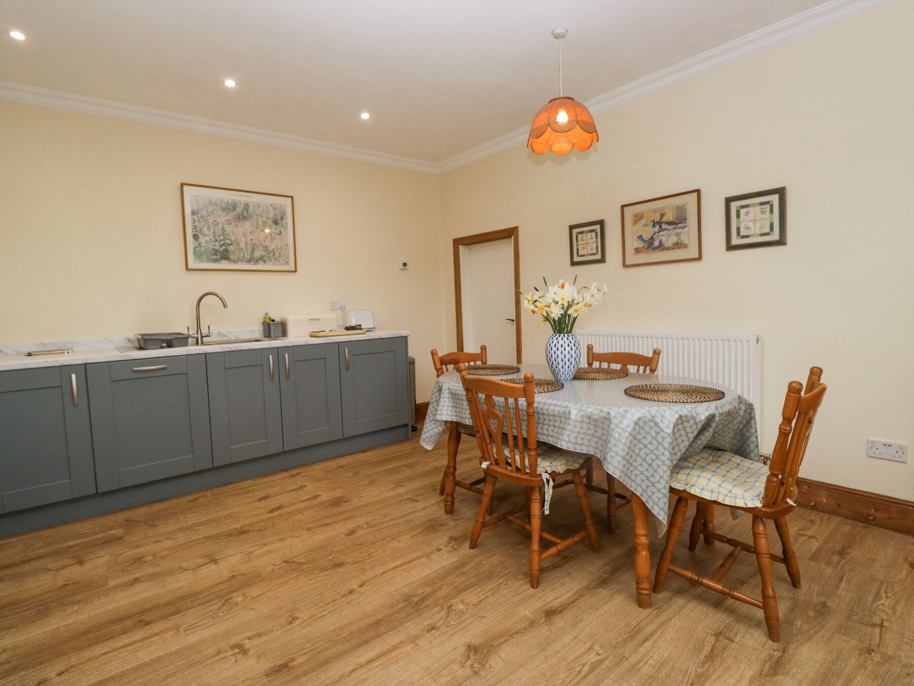 A dining area with a table set for four, surrounded by wooden chairs. A vase with flowers is on the table, and there are framed pictures on the wall.