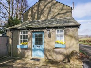A small stone cottage with a blue door and windows, a stone chimney, and a stone wall with a gravestone. - Winn's Cottage (Penrith)