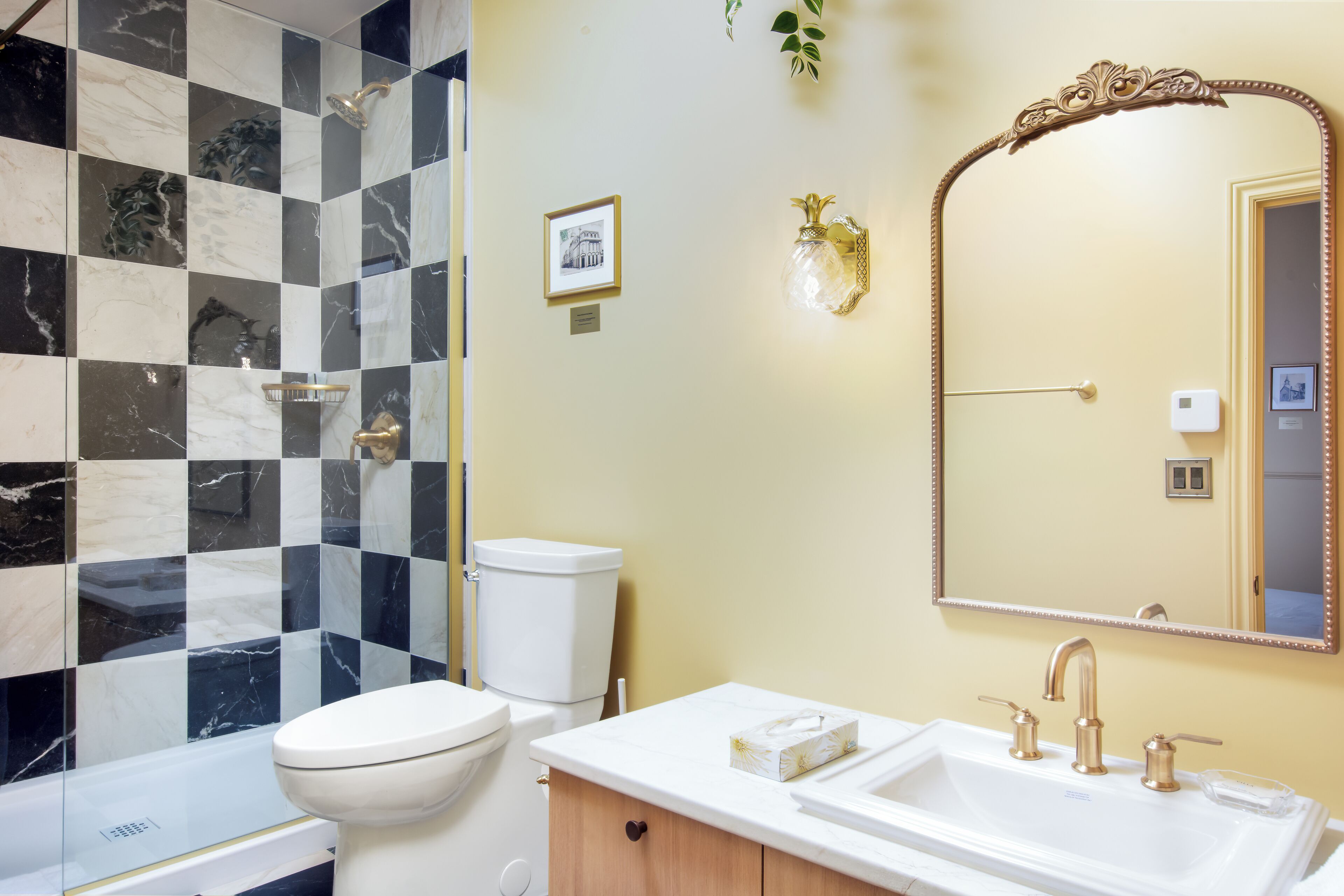 Um banheiro com parede de azulejos xadrez, vaso sanitário branco, pia com detalhes dourados e espelho com moldura ornamentada.