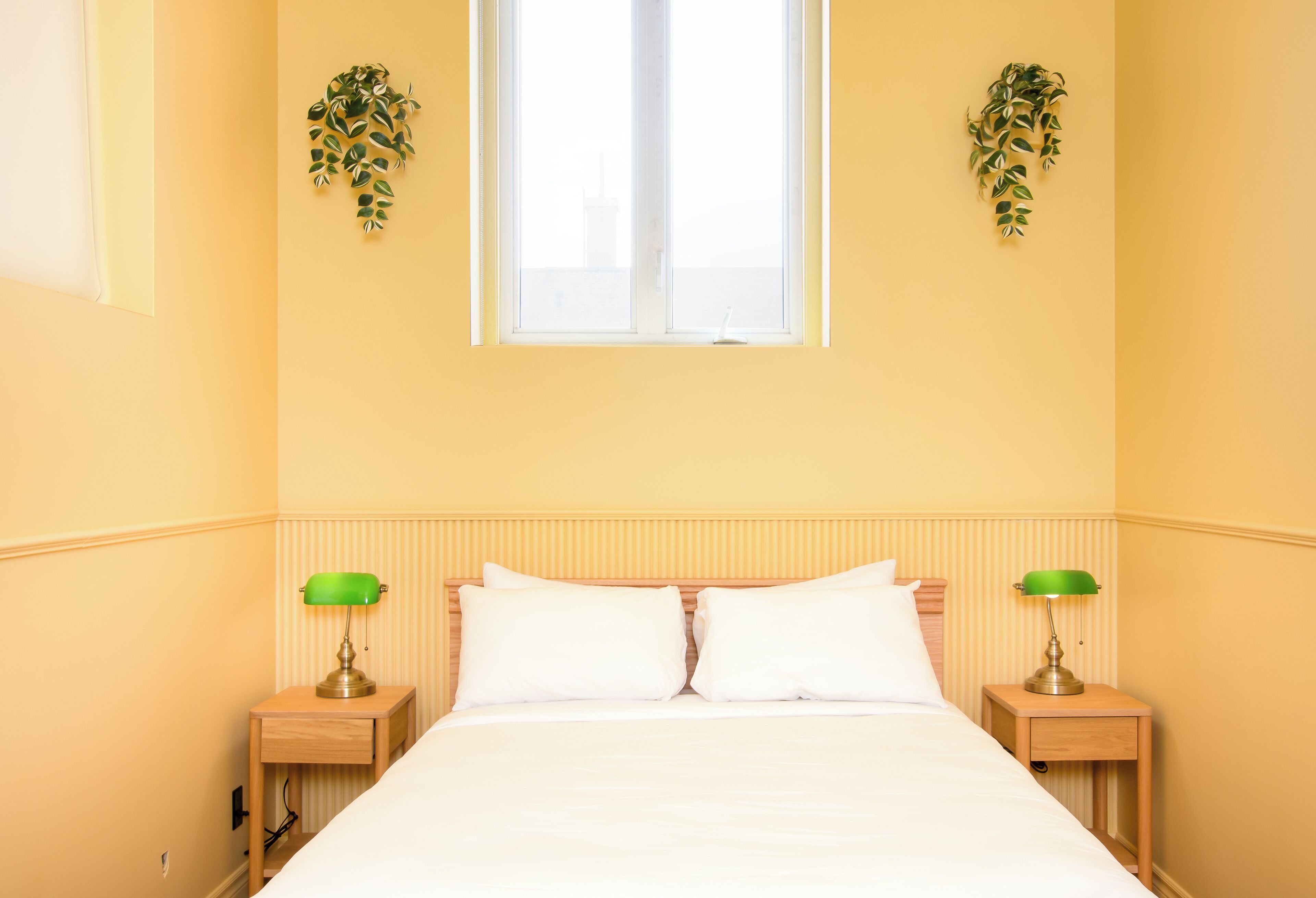 A hotel room with a bed, two bedside tables with lamps, a window, and two wall-mounted plants.