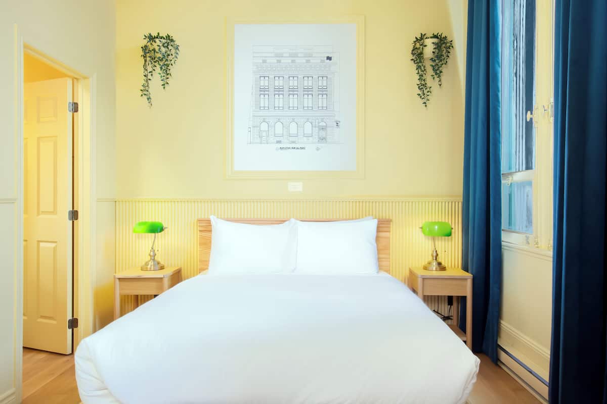 Una habitación de hotel con una cama grande, dos mesitas de noche con lámparas, un dibujo arquitectónico enmarcado en la pared y una ventana con cortinas azules.