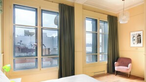 Une chambre d’hôtel avec de grandes fenêtres, un lit, une chaise et un bureau avec une lampe.