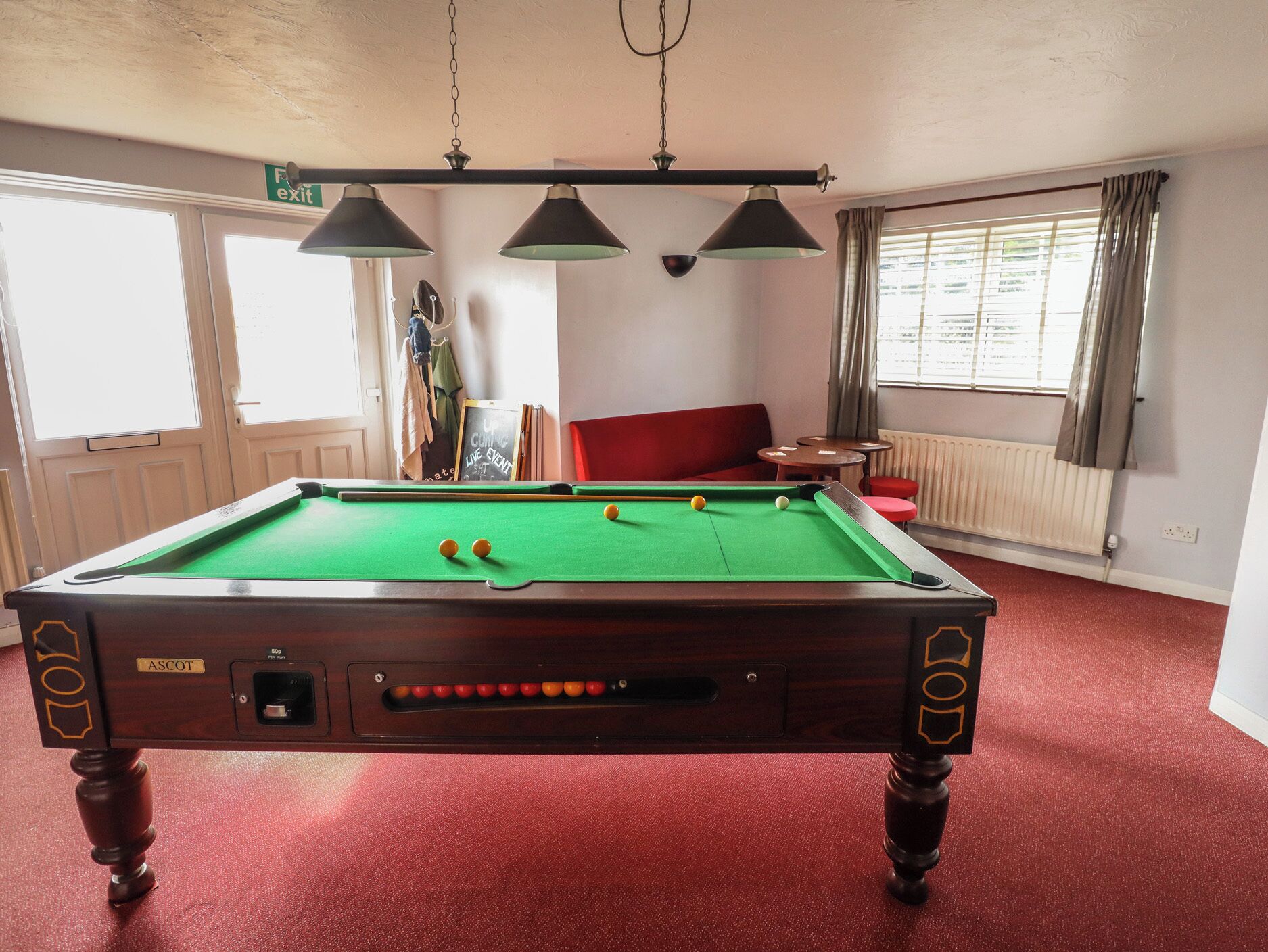 A pool table with a red carpet, a red sofa, and a window with curtains.