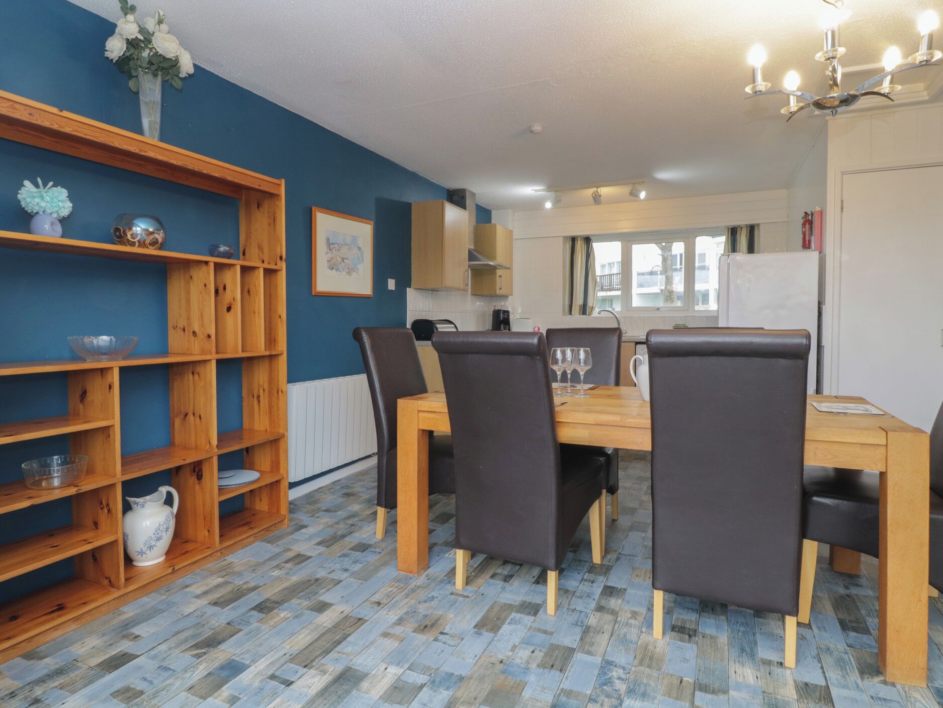 Un coin repas avec une table et des chaises en bois, une bibliothèque et, au fond, une cuisine.