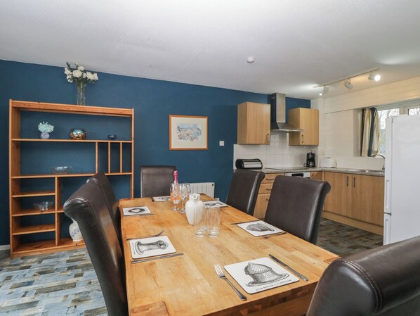 A dining room with a wooden table set for four, surrounded by leather chairs. A wooden bookshelf with decorative items is to the left.