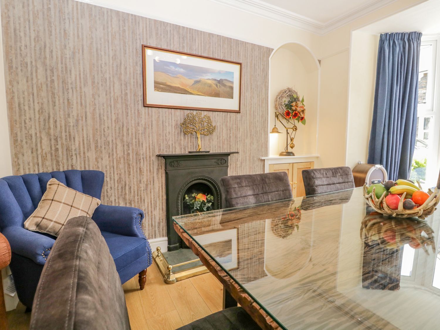 A living room with a blue sofa, a fireplace, a glass coffee table, and a dining table with a fruit basket.