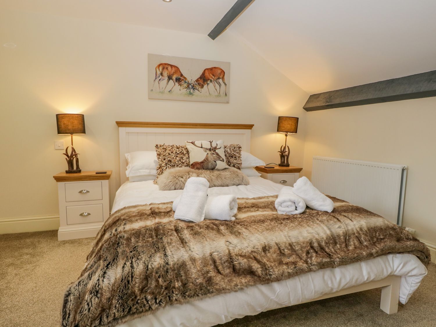 A bedroom with a large bed, two bedside tables, a headboard, and a painting of deer on the wall.