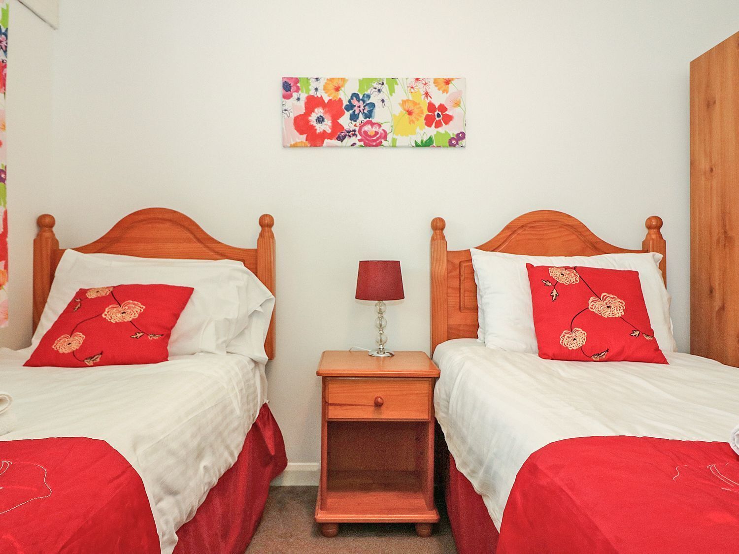 Quarto com duas camas de madeira, cada uma com roupa de cama branca e almofadas estampadas com flores vermelhas. Um criado-mudo com luminária e pequena gaveta fica entre as camas. Uma obra de arte floral colorida está pendurada na parede acima das camas.