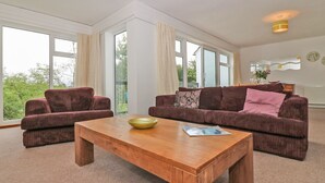 A spacious living room with two sofas, a wooden coffee table, and large windows offering a view of greenery. - Tamar Cottages 35 (Gunnislake)