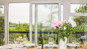 A dining table set for six with a bottle of wine, a vase of flowers, and a view of greenery through large windows. - Tamar Cottages 35 (Gunnislake)