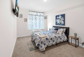 A bedroom with a bed, a window with curtains, a painting on the wall, and a small bedside table. - Blue Bliss 2BR 2BTH with FREE Parking, Near NYC & EWR Airport (East Orange)