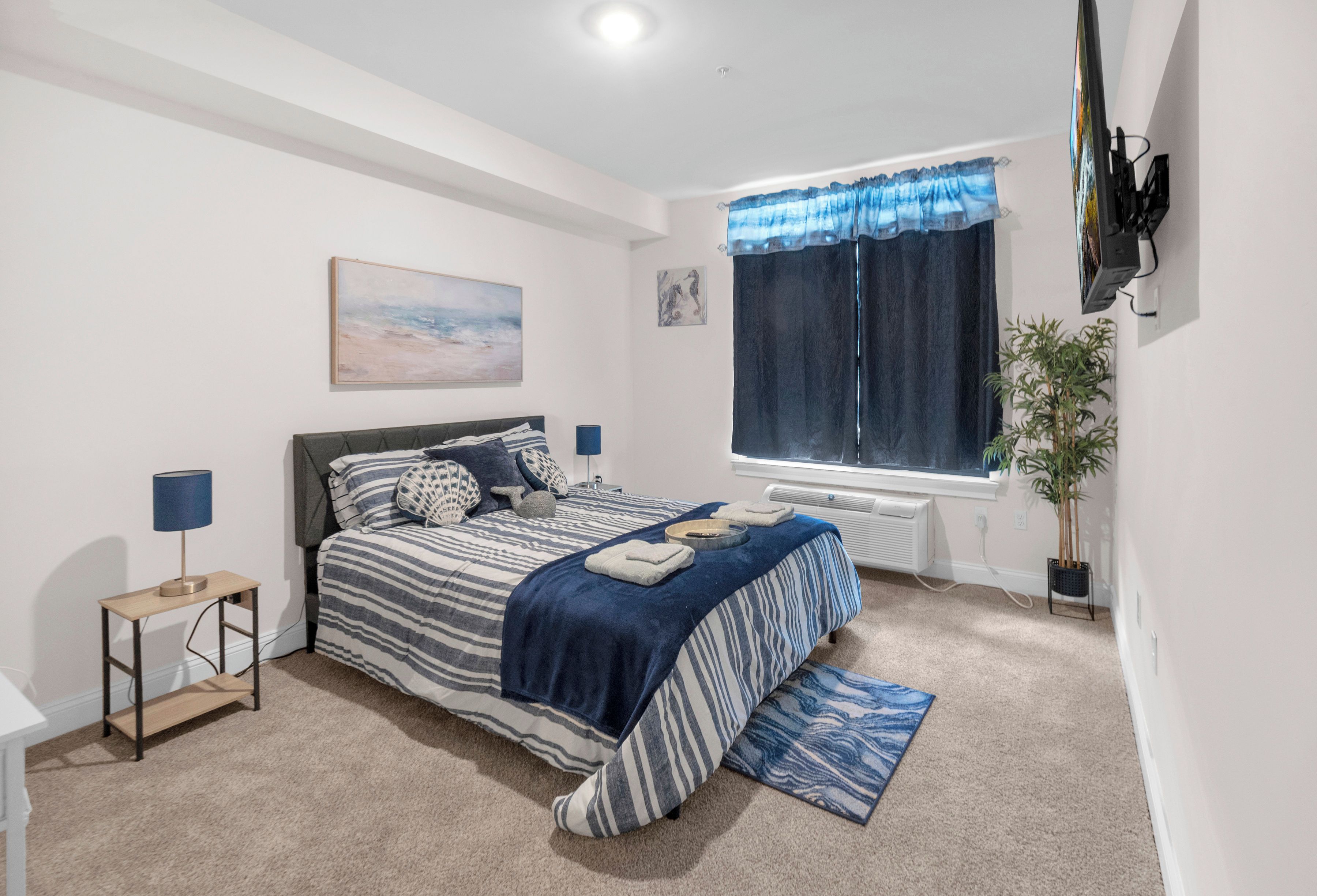 A bedroom with a bed, bedside tables, a window with curtains, a radiator, a plant, and artwork on the wall.