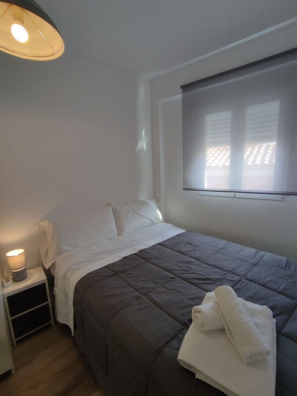A neatly made bed with a grey duvet, white pillows, and a white blanket. There is a bedside table with a lamp and a small white object. A window with blinds is visible in the background.