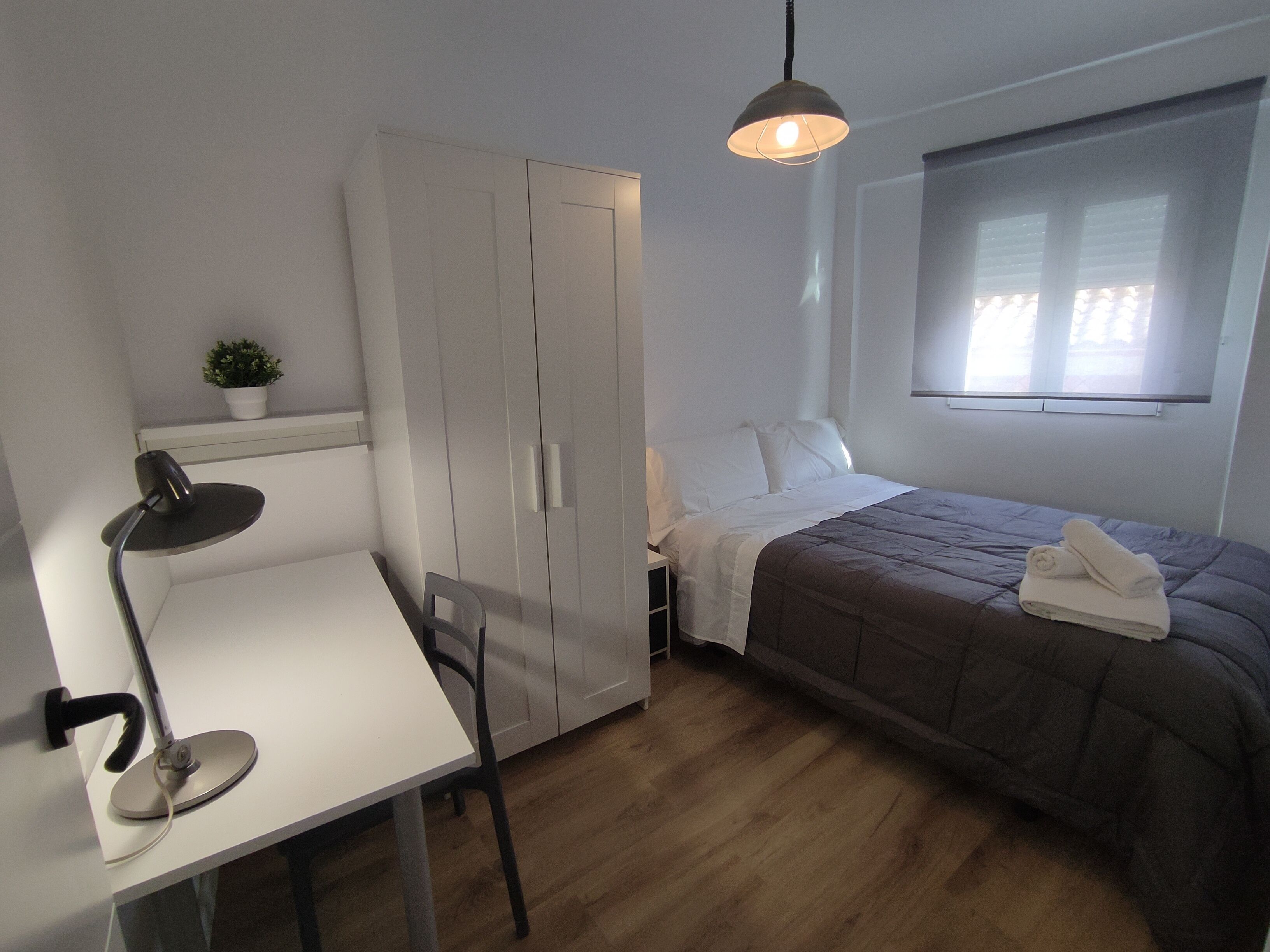 A bedroom with a bed, a desk, a chair, a wardrobe, and a hanging light fixture.