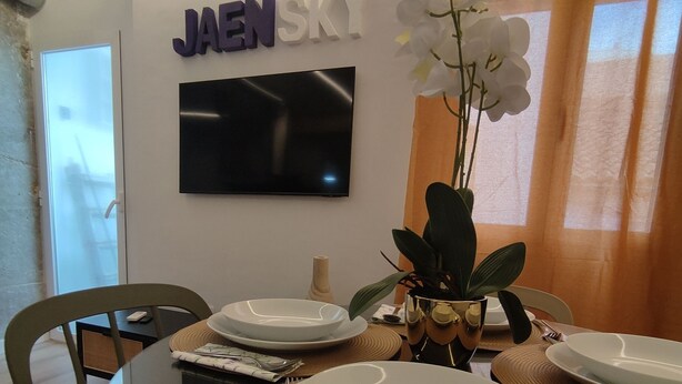 Un comedor con una mesa de vidrio, platos blancos y un jarrón con flores blancas. Un televisor de pantalla plana está montado en la pared, y hay un letrero con el texto 
