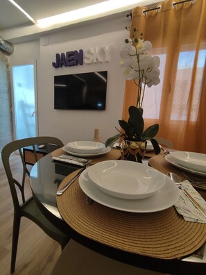 Un comedor con una mesa de vidrio, platos blancos y un jarrón con flores blancas. Un televisor de pantalla plana está montado en la pared, y hay un letrero con el texto 