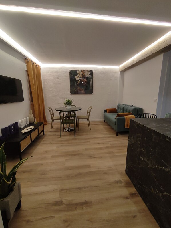 Una sala de estar moderna con una televisión de pantalla plana, una mesa de comedor con sillas, un sofá y una mesa de centro con una planta.