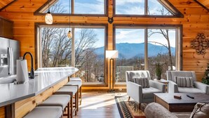 Une cabane en bois comprenant une cuisine, un coin repas et un espace détente près d’une grande fenêtre donnant sur les montagnes.