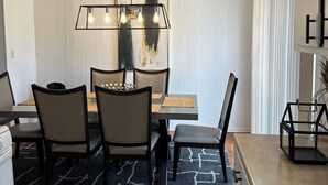 Ett modernt matområde med ett rektangulärt bord, fyra stolar, en hänglampa med synliga glödlampor, en stor abstrakt målning och en köksbänk med en diskho.