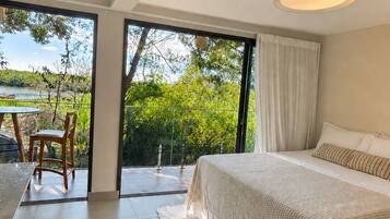 A modern bedroom with a large bed, a view of greenery, and a dining area with a table and chairs.