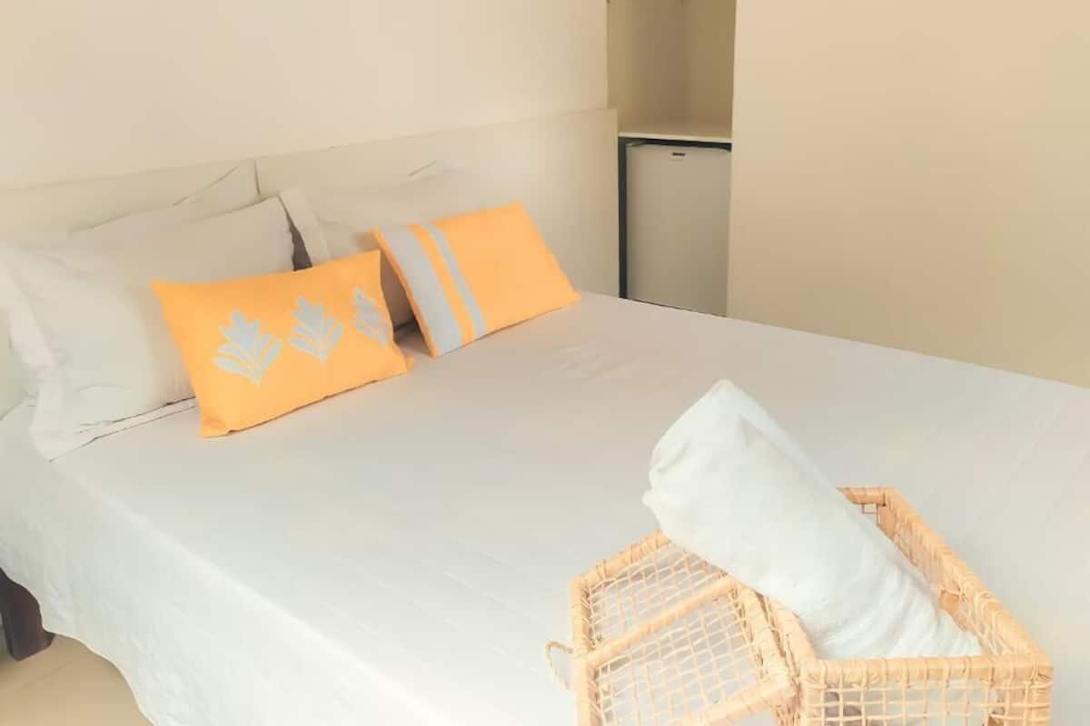 A neatly made bed with white and orange pillows, a white blanket, and a wicker basket with a white pillow.