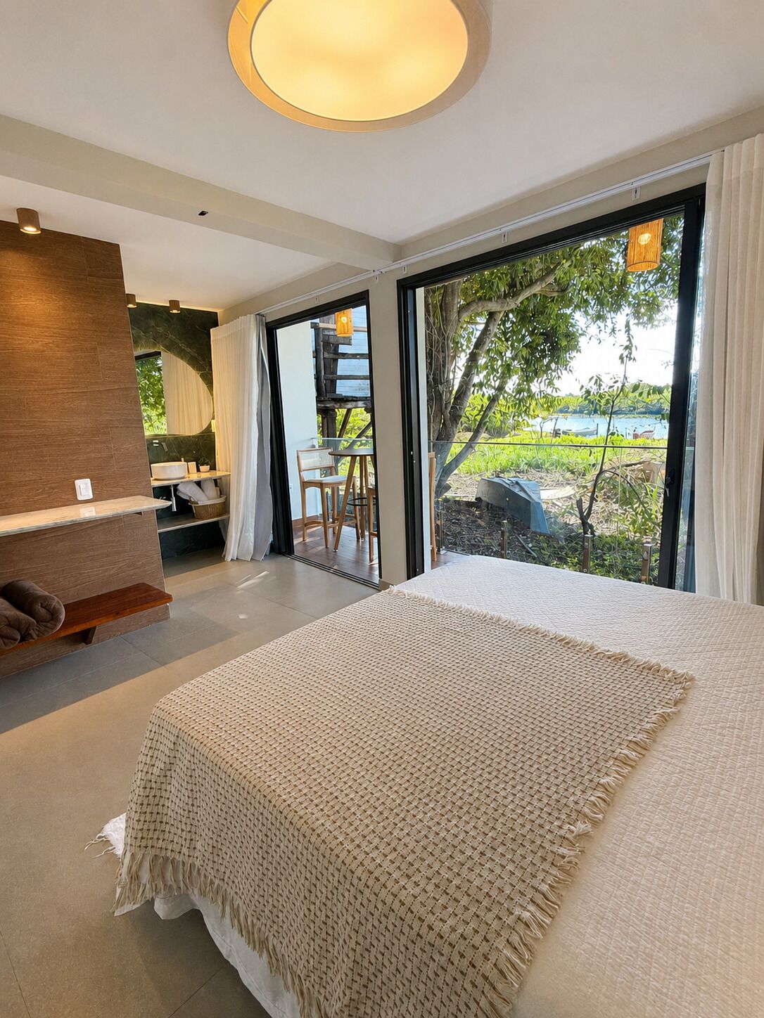 A modern bedroom with a large bed, a seating area with a table and chairs, and a view of a natural landscape through large windows.