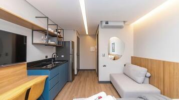 A compact kitchenette with a fridge, sink, and microwave, a bed with a headboard, and a small bedside table.