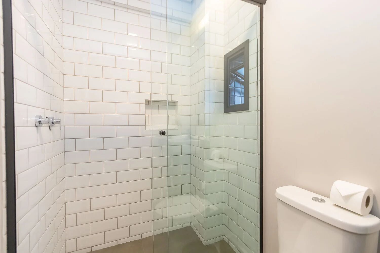 Um chuveiro com paredes de vidro, azulejos brancos tipo metrô, uma pequena janela e um rolo de papel higiênico.