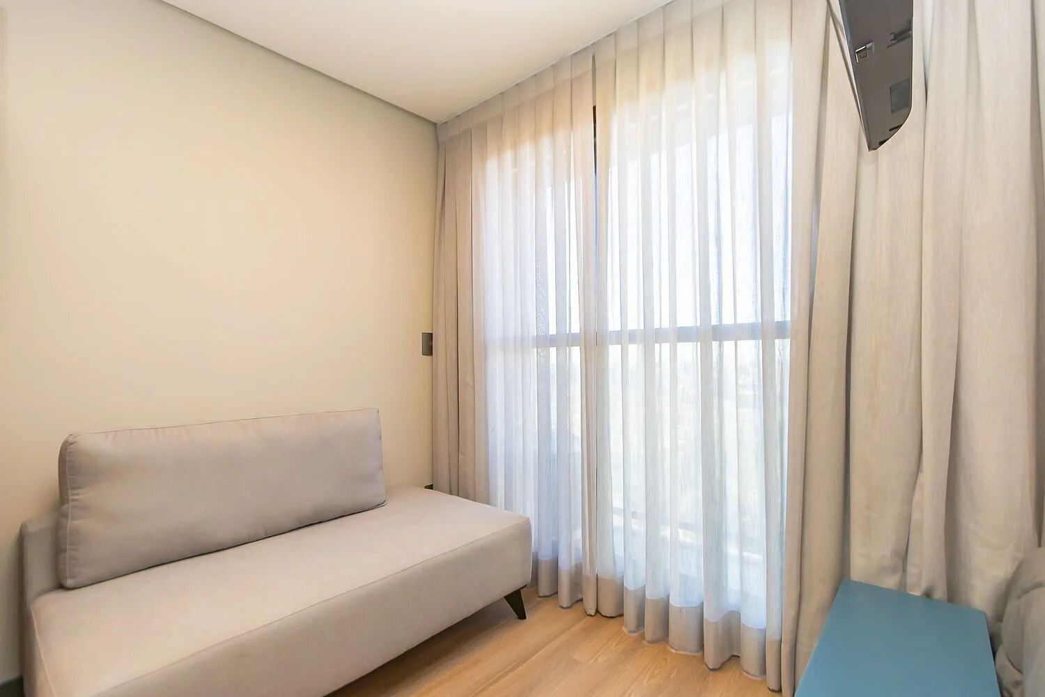 Une pièce au décor minimaliste, avec un canapé de couleur claire, une grande fenêtre ornée de rideaux légers et une table bleue.